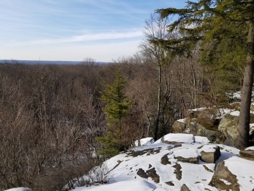 A snowy landscape with rocky outcrops and bare trees, overlooking a distant forested valley under a clear sky.
