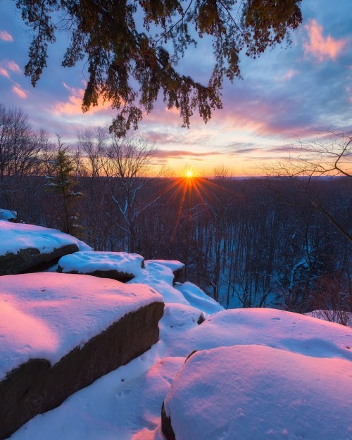 Sunset over a snowy landscape, with rocks in the foreground and colorful clouds in the sky.