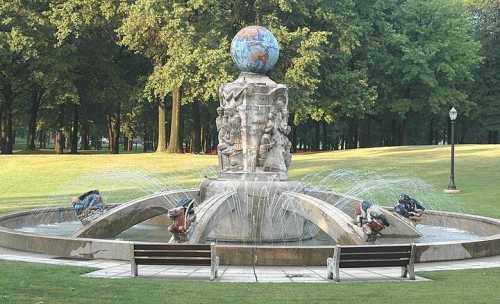 A decorative fountain with a globe on top, surrounded by sculptures and water jets, set in a green park.