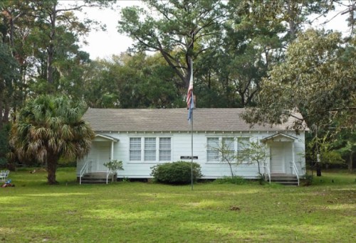 A white, single-story house surrounded by trees and grass, featuring an American flag in front.