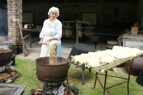 A woman in historical attire stirs a pot over a fire, with fabric drying on a rack nearby in a rustic setting.