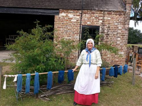 A woman in historical attire stands beside a line of indigo-dyed fabric hanging outside a brick building.