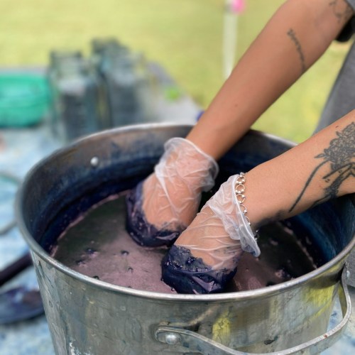 A person wearing gloves is mixing dye in a large metal bucket, with hands submerged in a purple liquid.