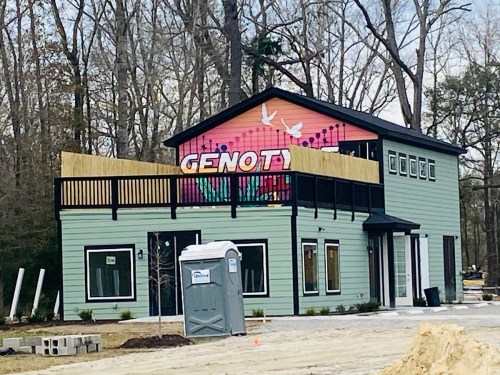 A two-story building with a colorful mural reading "GENOT" and a wooden deck, surrounded by trees and construction materials.