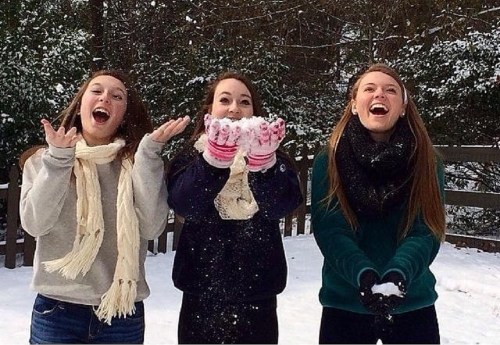 Three friends joyfully play in the snow, tossing snowflakes in the air while wearing winter scarves and gloves.