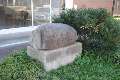 A large stone monument inscribed with "Aaron Burr Rock," surrounded by greenery and a building in the background.