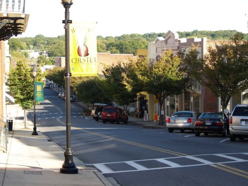 A quiet street in Chester, lined with trees and shops, featuring a welcome banner and parked cars.