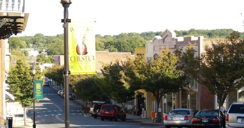 A quiet street in Chester, lined with trees and shops, featuring a welcome banner and parked cars.