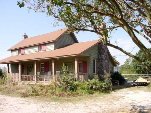 A two-story house with a brown roof, green siding, and red shutters, surrounded by trees and a gravel path.