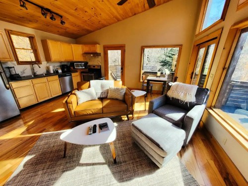 Cozy living space with a kitchen, wooden accents, large windows, and comfortable seating in a sunlit room.