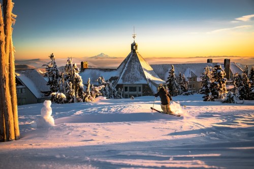 A skier glides through fresh snow with a mountain backdrop and a snowman nearby, illuminated by a golden sunrise.