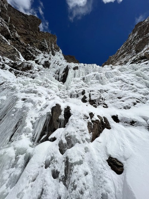 A frozen waterfall cascading down rocky cliffs under a bright blue sky with scattered clouds.