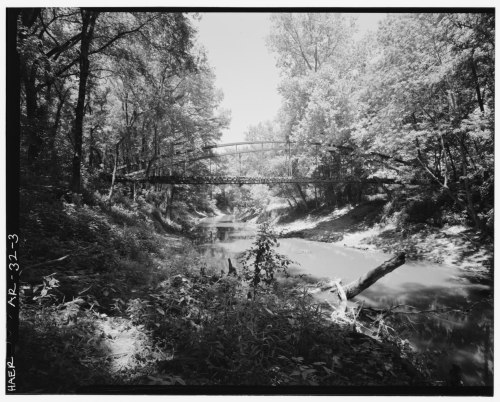 A black and white photo of a bridge over a calm river, surrounded by lush trees and greenery.