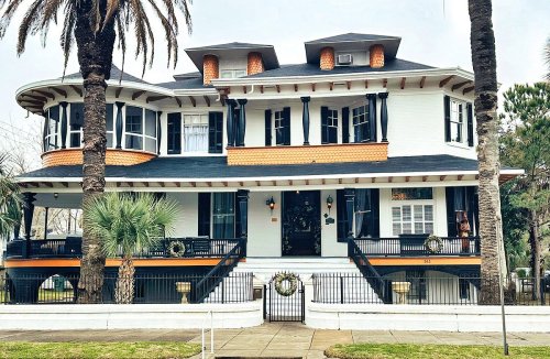 A large, two-story house with a wraparound porch, palm trees, and festive decorations on the balcony.