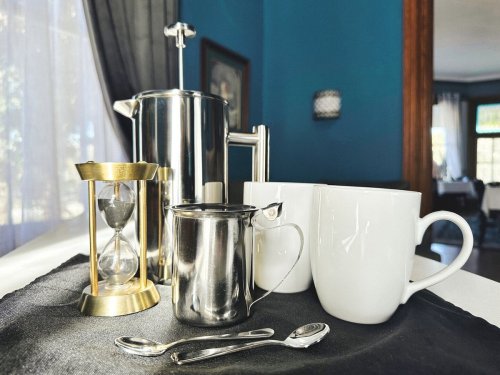 A coffee setup featuring a French press, two white mugs, a milk pitcher, an hourglass, and silver spoons on a black cloth.