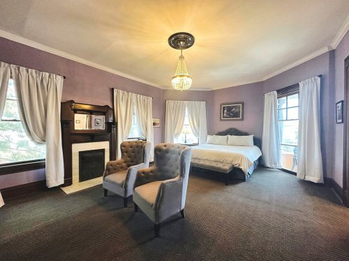 A cozy bedroom with a bed, two armchairs, a fireplace, and large windows, featuring soft purple walls and elegant decor.