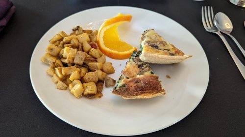 A plate with two slices of quiche, roasted potatoes, and a slice of orange on a black table.