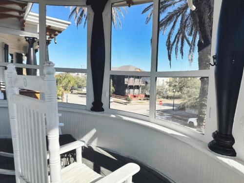 A cozy porch with white rocking chairs, palm trees outside, and a view of a quiet street and nearby buildings.