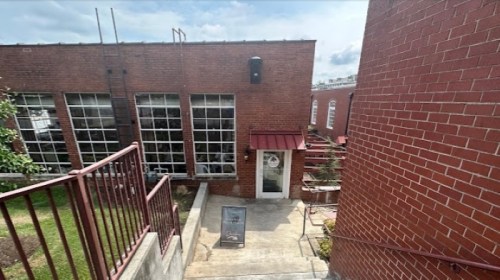 A brick building with large windows, a red awning, and stairs leading down to a door. Greenery is visible nearby.