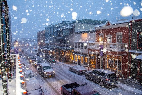 A snowy street scene with cars and shops, illuminated by warm lights, as large snowflakes fall heavily.