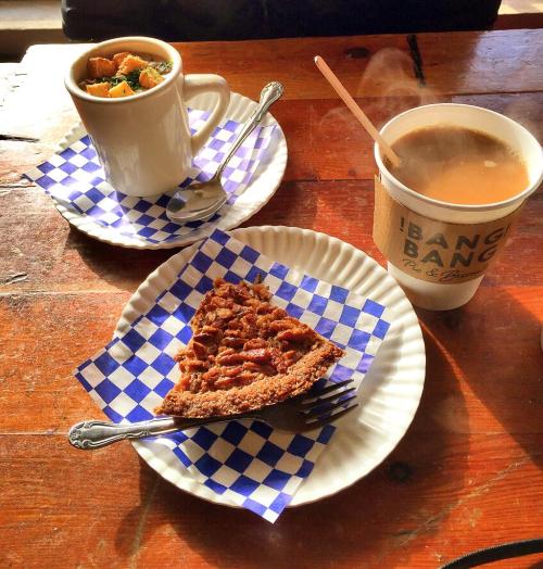 A slice of pecan pie on a blue checkered plate, with a cup of soup and coffee in the background on a wooden table.