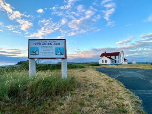 Sign for Point Wilson Light with a historic lighthouse and grassy area under a blue sky with scattered clouds.