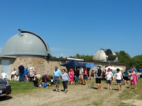 A crowd of people gathers outside a building with two domed observatories under a clear blue sky.