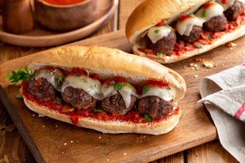 Two meatball subs with marinara sauce and melted cheese on a wooden cutting board, garnished with parsley.