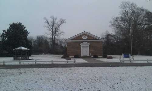 A brick building with a white door, surrounded by snow, trees, and a gazebo in a winter landscape.