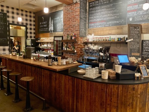 A cozy café interior featuring a wooden bar, coffee equipment, and a chalkboard menu against a brick wall.