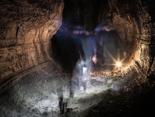 A dimly lit cave with blurred figures walking through, illuminated by a single light source.