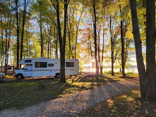 A white RV parked among colorful autumn trees, with sunlight streaming through the leaves and a lake in the background.