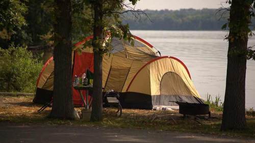 A colorful camping tent by a calm lake, surrounded by trees and a small table with camping gear nearby.