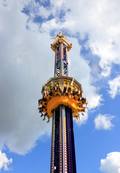 A tall amusement park ride with a colorful design, featuring riders suspended at the top against a blue sky with clouds.