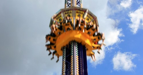 A thrilling amusement park ride spins high against a blue sky, with excited riders hanging on.