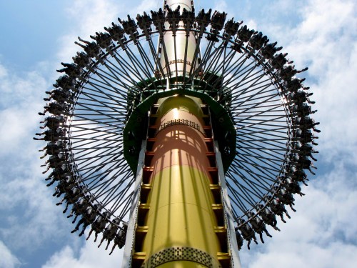 A tall amusement park ride with riders suspended in a circular formation against a blue sky with clouds.