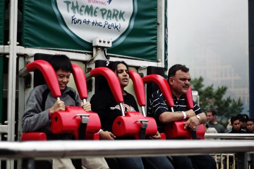 Three people seated on a thrill ride, secured with red harnesses, looking apprehensive before the drop.