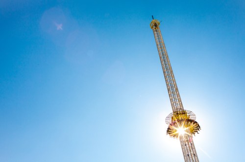 A tall amusement park ride against a clear blue sky, with sunlight creating a starburst effect at the base.