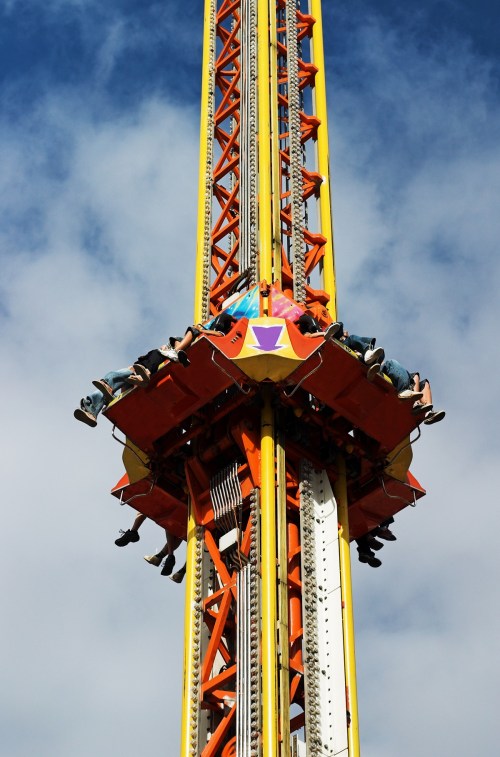 A tall amusement park ride with passengers seated at the top, ready to drop against a blue sky with clouds.