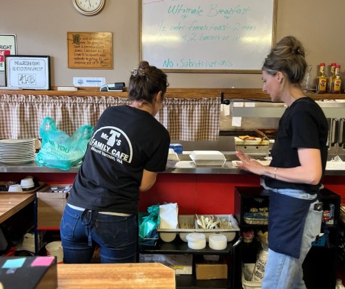 Two women working in a café kitchen, preparing food and organizing supplies at the counter.