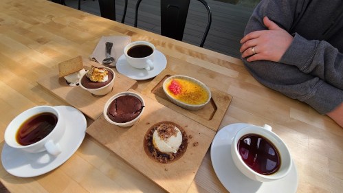 A wooden table with two cups of coffee, desserts including a tart, crème brûlée, and a chocolate treat, and a hand.