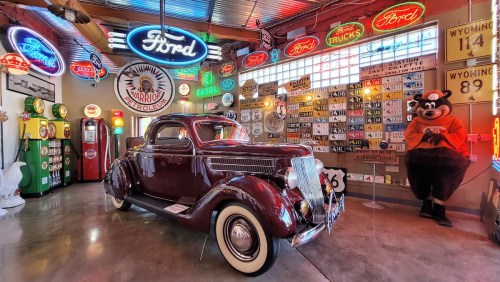 A vintage maroon car in a colorful garage filled with neon signs, gas pumps, and memorabilia.