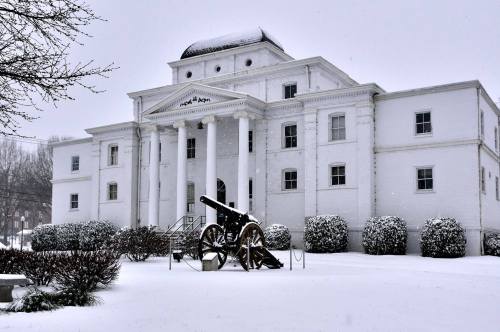 A white building with columns and a cannon in front, surrounded by snow-covered ground and trees.