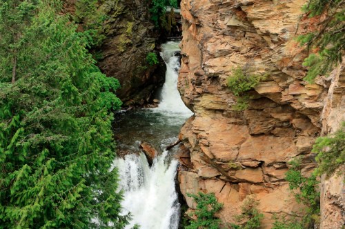 A waterfall cascades between rocky cliffs, surrounded by lush green trees in a serene natural setting.
