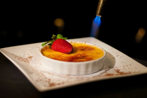 A creamy crème brûlée topped with a fresh strawberry, being caramelized with a kitchen torch on a white plate.