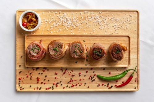 A wooden platter with five meat rolls, garnished with herbs, surrounded by spices and a small bowl of sauce.