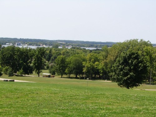 A scenic view of a green park with trees, a grassy field, and a distant lake under a clear blue sky.