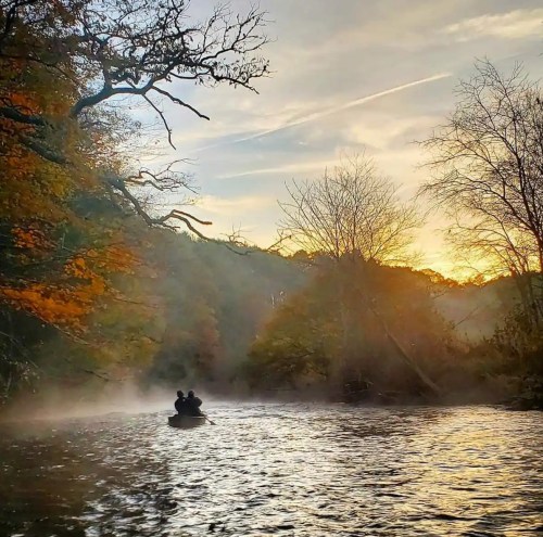 A serene river scene at sunset, with two people in a canoe surrounded by mist and autumn foliage.