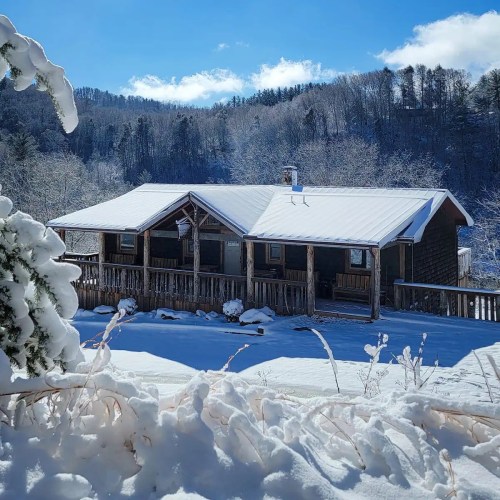A cozy wooden cabin covered in snow, surrounded by trees and mountains under a clear blue sky.