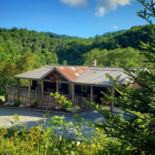 A rustic cabin surrounded by lush green trees and mountains under a blue sky with fluffy clouds.